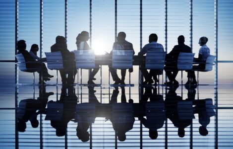 Executives In Silhouette Meeting Around A Boardroom Table In A Sunny Office With Many Windows And Blue Sky.