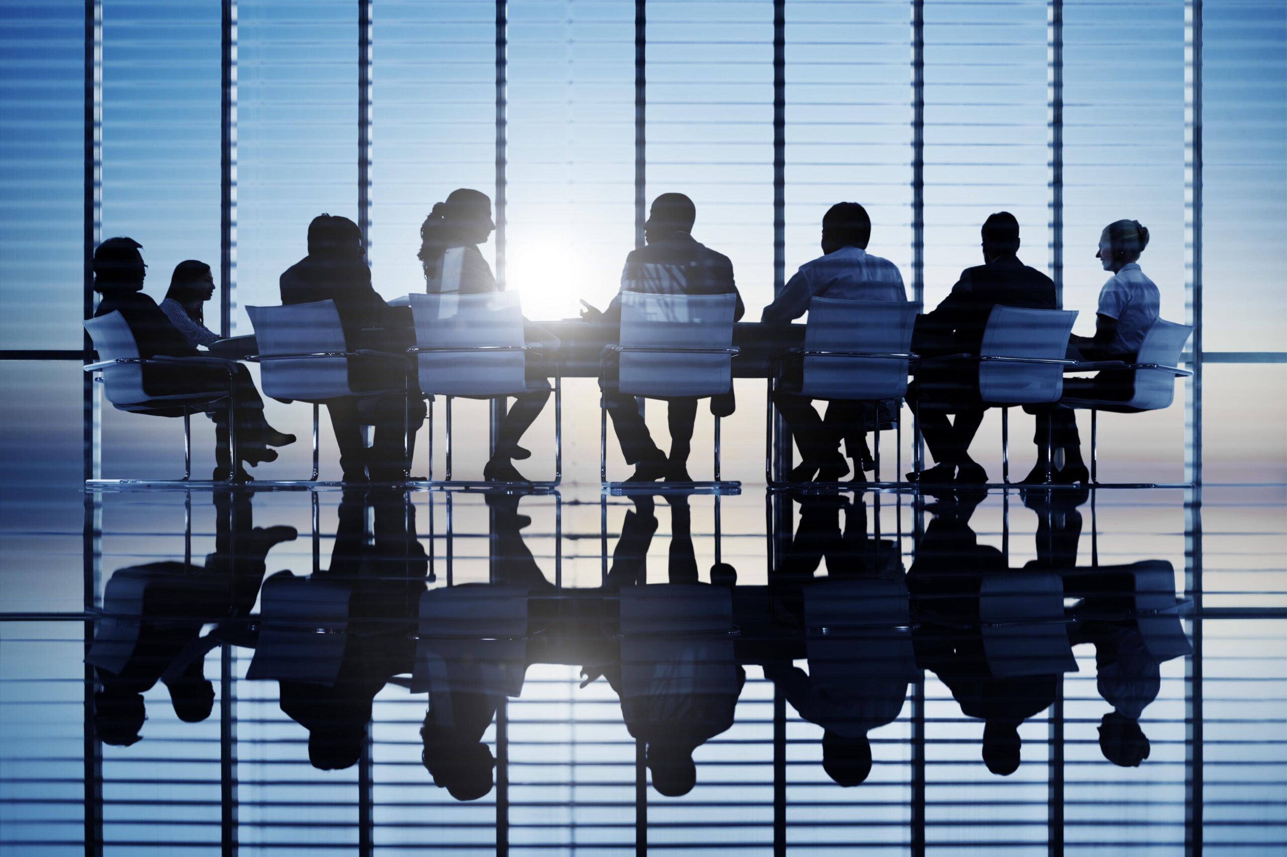 Executives in silhouette meeting around a boardroom table in a sunny office with many windows and blue sky.