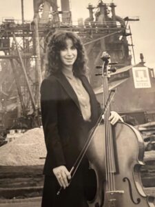 Jill Harmon, black and white photo from 1980s, standing in business suit outside a steel mill, holding a cello and bow. 