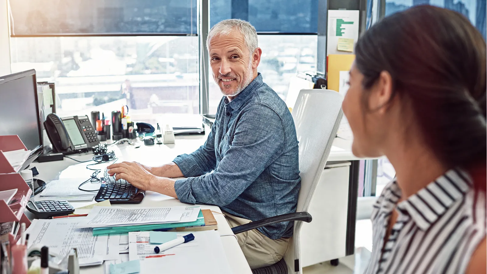 A mature worker with gray hair at a computer chats with a younger coworker.