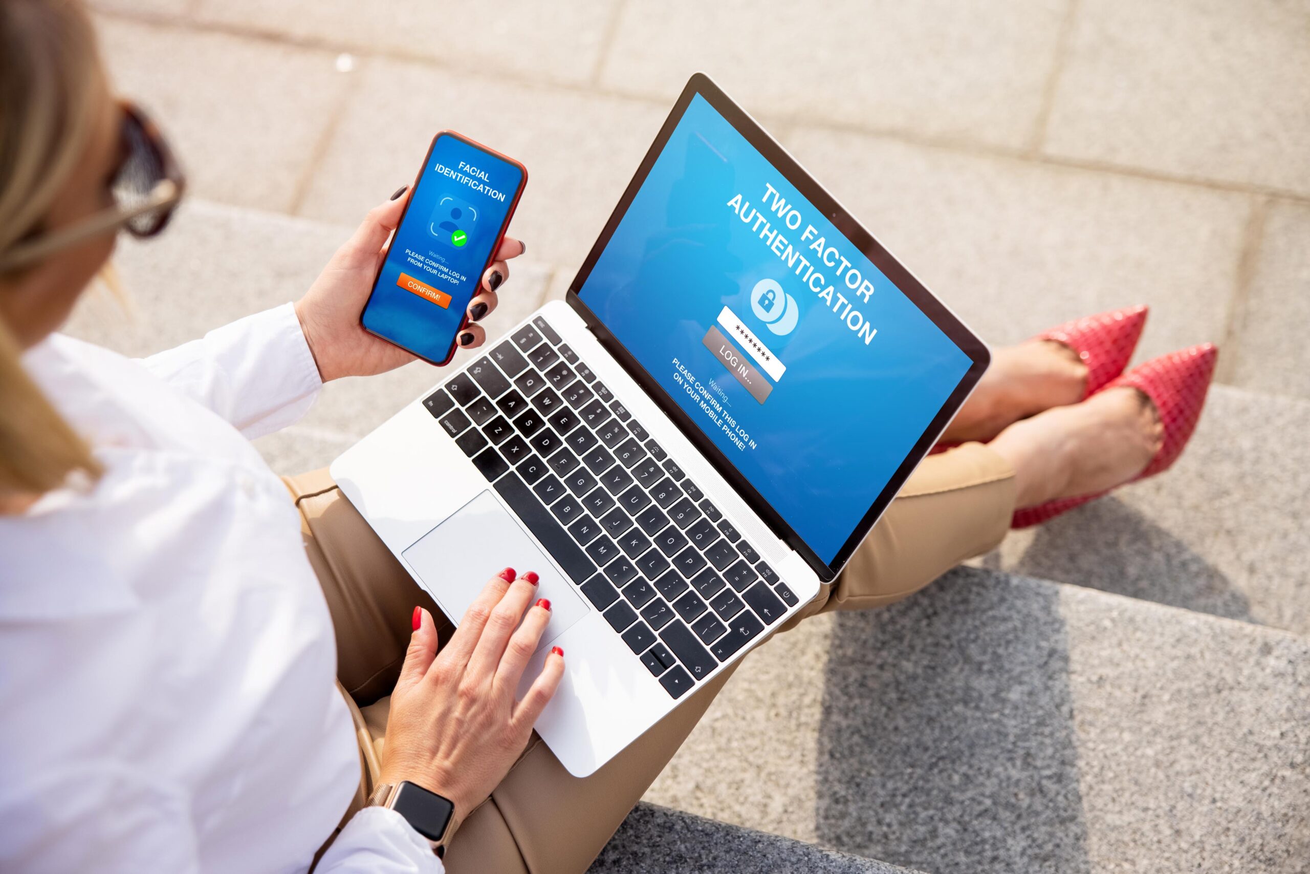 Women in business attire white shirt tan pants holding laptop and smartphone, sitting outside on steps.